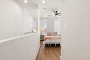 Ein schmaler Flur führt zu einem Schlafzimmer mit einem Bett, einem Deckenventilator und einem Fenster mit Jalousien.