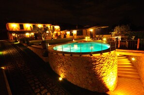 A nighttime view of a pool area with a stone wall surrounding it, a staircase leading up to a covered deck, and a building with multiple windows and lights in the background. - La Valle Dei Casali B&B (Rome)
