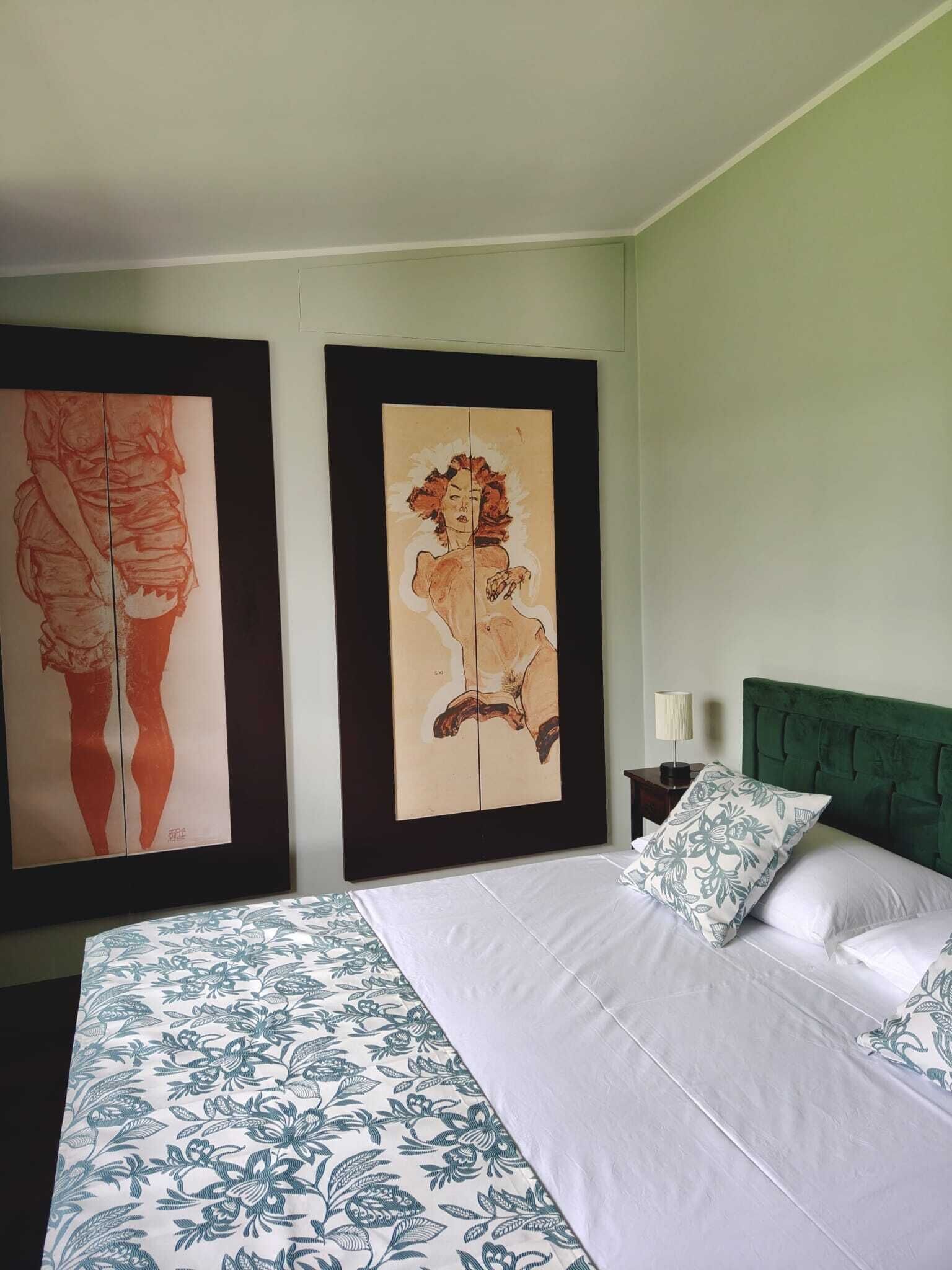 A hotel room with a bed, two framed artworks, a green headboard, and a nightstand with a lamp.