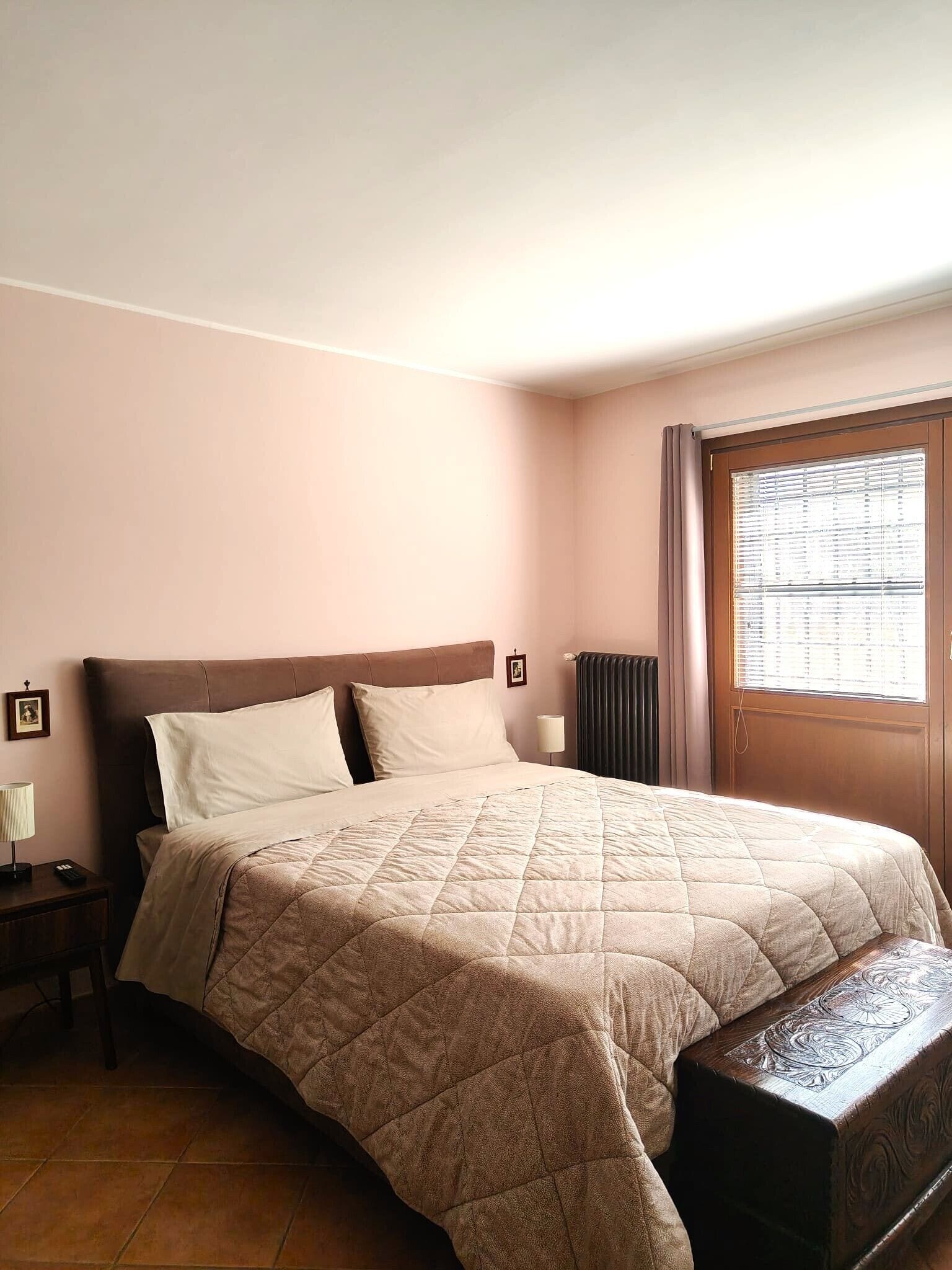 A bedroom with a large bed, a nightstand, a lamp, a radiator, and a window with blinds.
