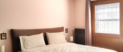 A bedroom with a large bed, a nightstand, a lamp, a radiator, and a window with blinds.