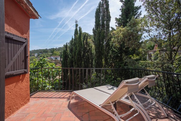Een balkon met een tafel en stoelen, met uitzicht op een tuin met hoge bomen en in de verte huizen.
