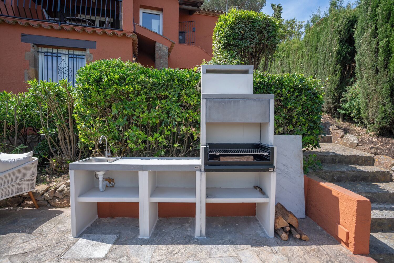 Eine Außenküche mit Spüle und Grill, umgeben von Steinstufen und Grünpflanzen.