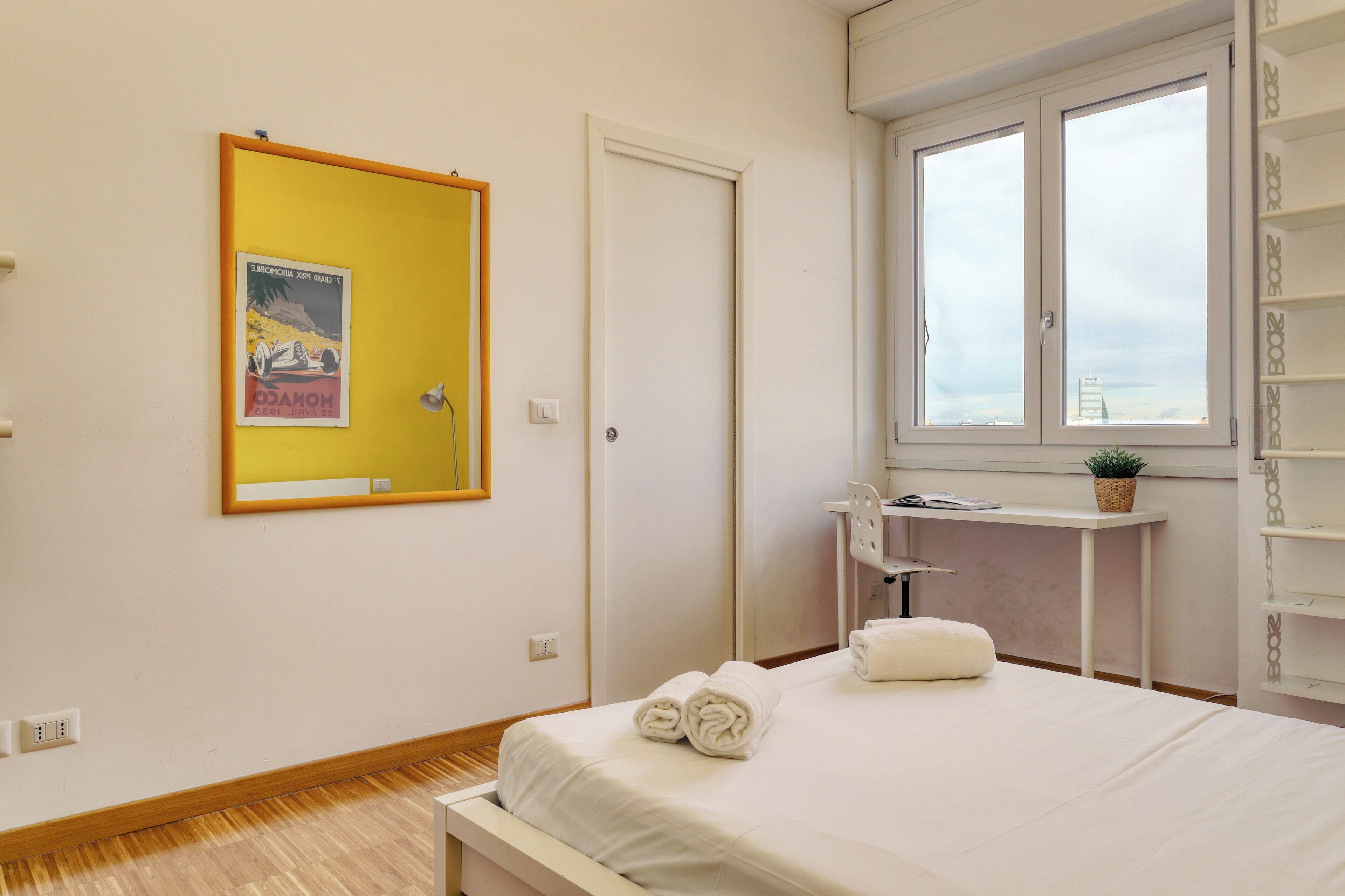 A hotel room with a bed, a small desk, a window, and a mirror on the wall.