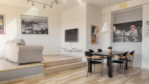 A modern living room with a sofa, a dining table with chairs, a TV mounted on the wall, and a staircase leading to an upper level. - Stylish 2BR flat next to Navigli (Milan)