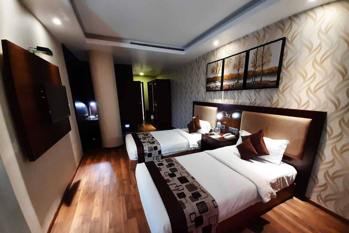 A hotel room with two beds, a flat-screen TV mounted on the wall, a bedside table with a lamp, and a patterned wallpaper.
