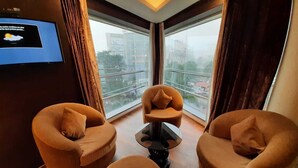 Una habitación de hotel moderna con dos sillones, una mesita de café y un televisor de pantalla plana montado en la pared. Amplios ventanales ofrecen vistas a la ciudad.