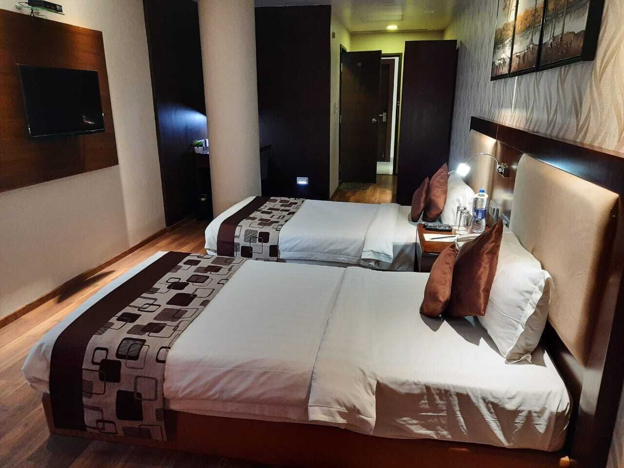 A hotel room with two beds, a TV mounted on the wall, and a bedside table with a lamp.