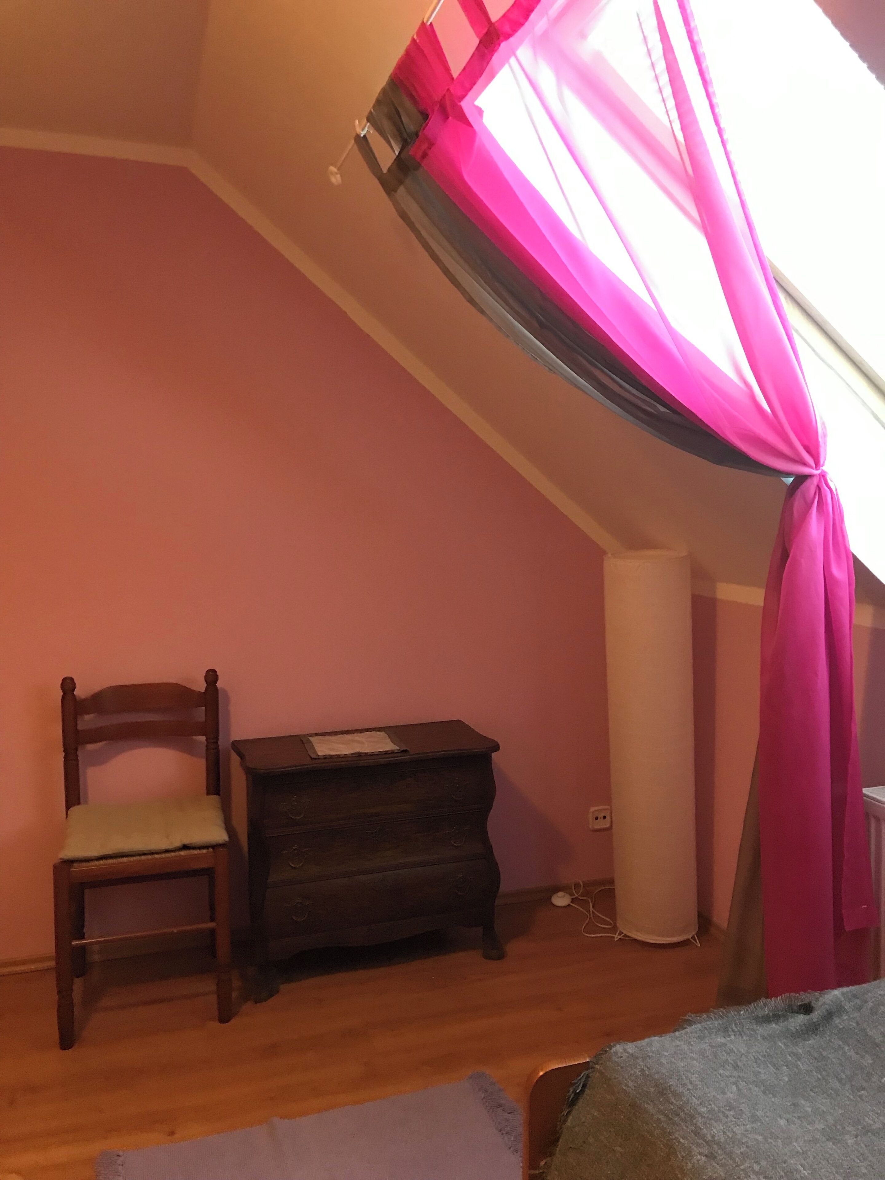 A room with a sloped ceiling, a wooden chair, a small wooden chest, and a pink curtain.