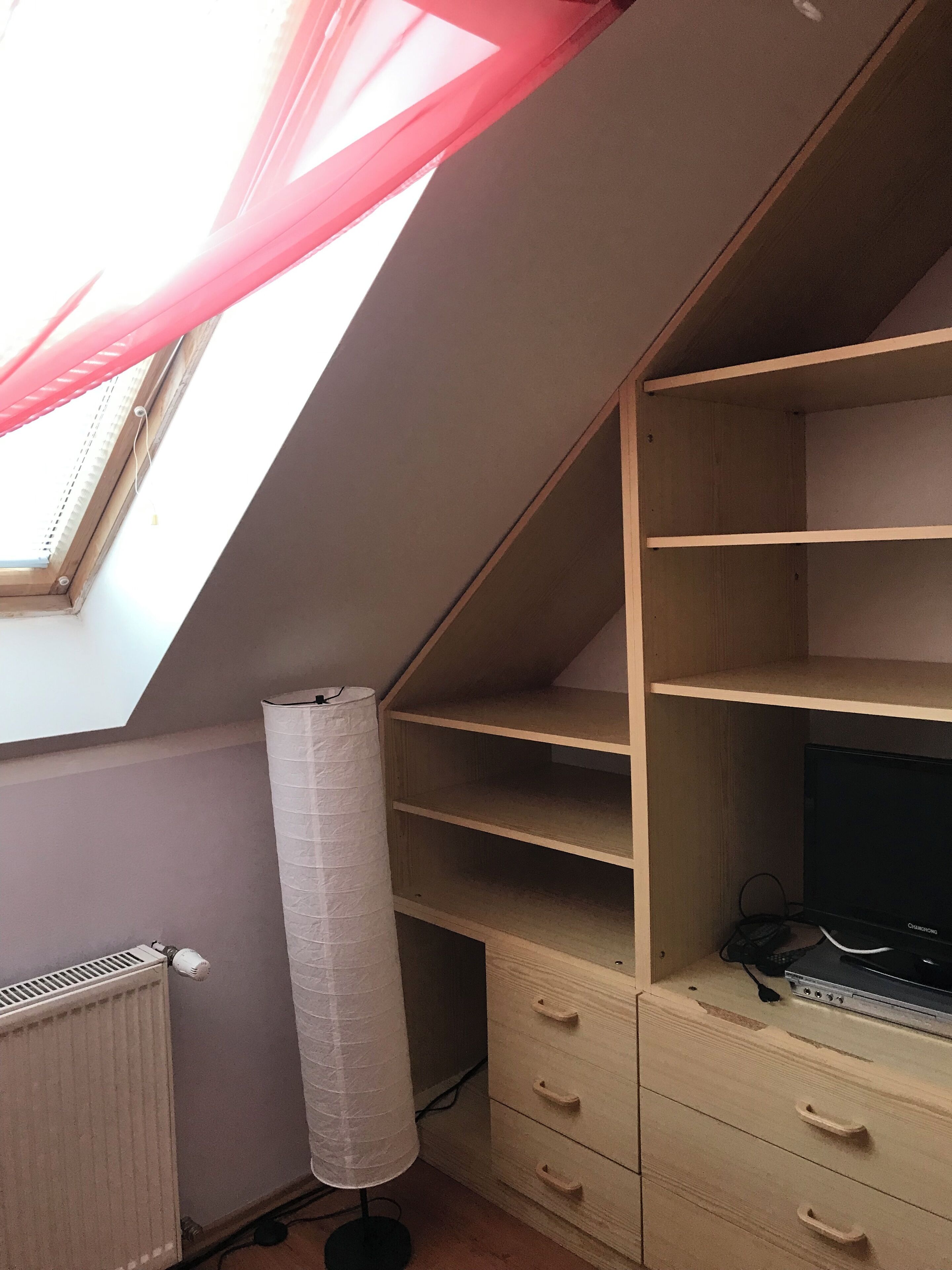 A room with a sloped ceiling, a skylight, a radiator, a lamp, and a wooden cabinet with drawers.