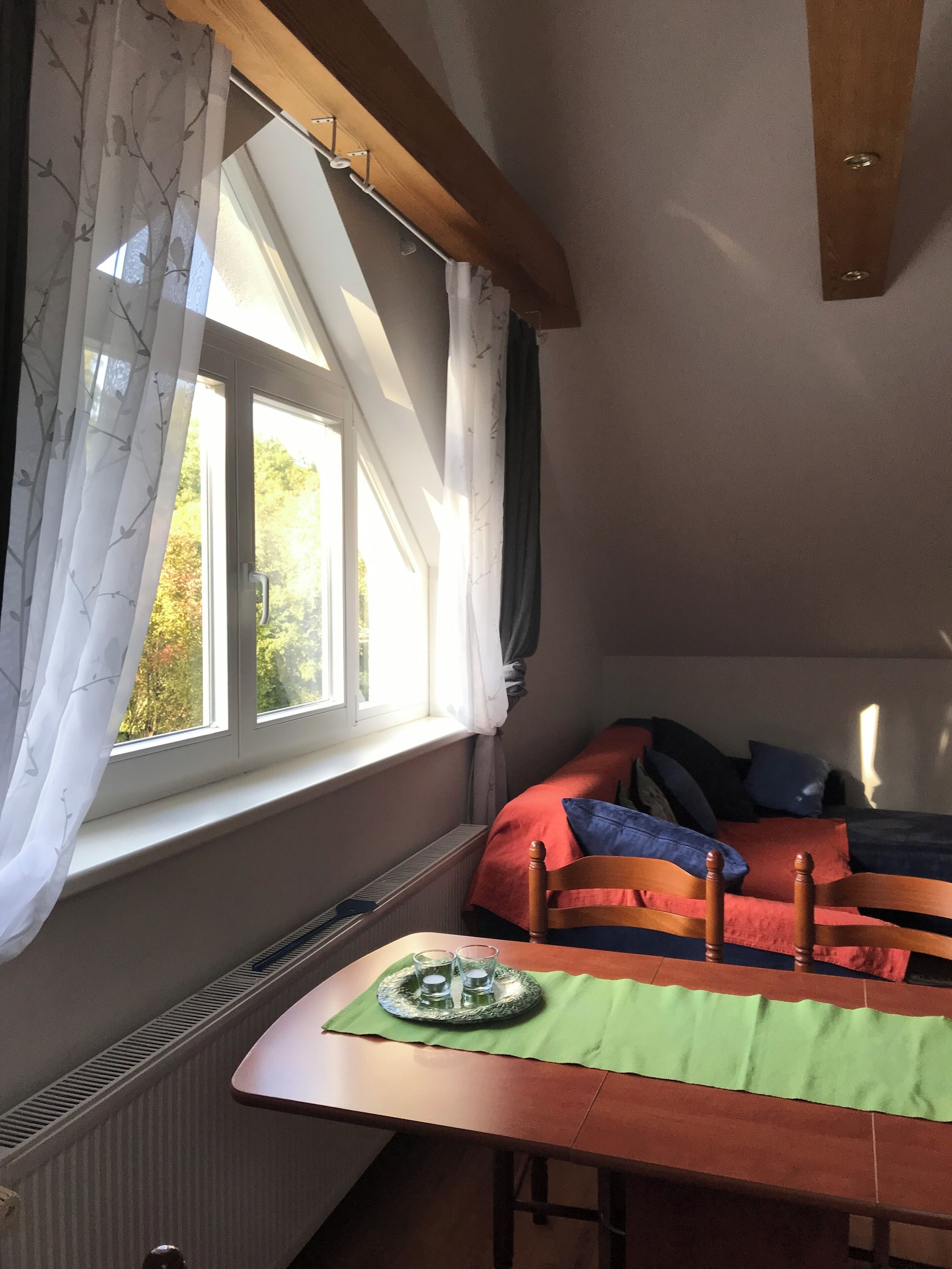 A room with a wooden table, a window with sheer curtains, a radiator, and a bed with a red and blue blanket.
