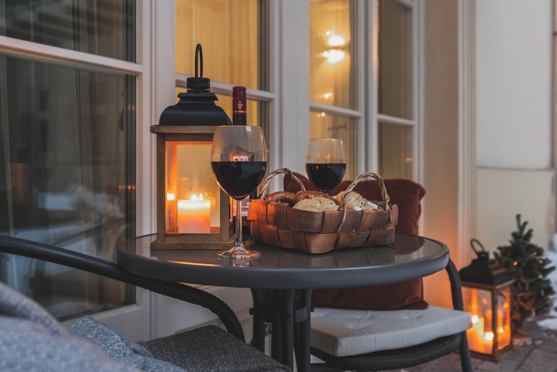 A round table with a candle, two glasses of red wine, a lantern, and a basket of bread on a balcony.
