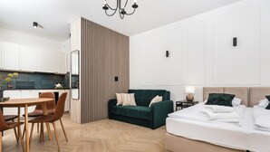 Ett modernt hotellrum med ett trägolv i fiskbensmönster, en grön soffa, en säng med vita sängkläder och ett litet sängbord med en lampa.