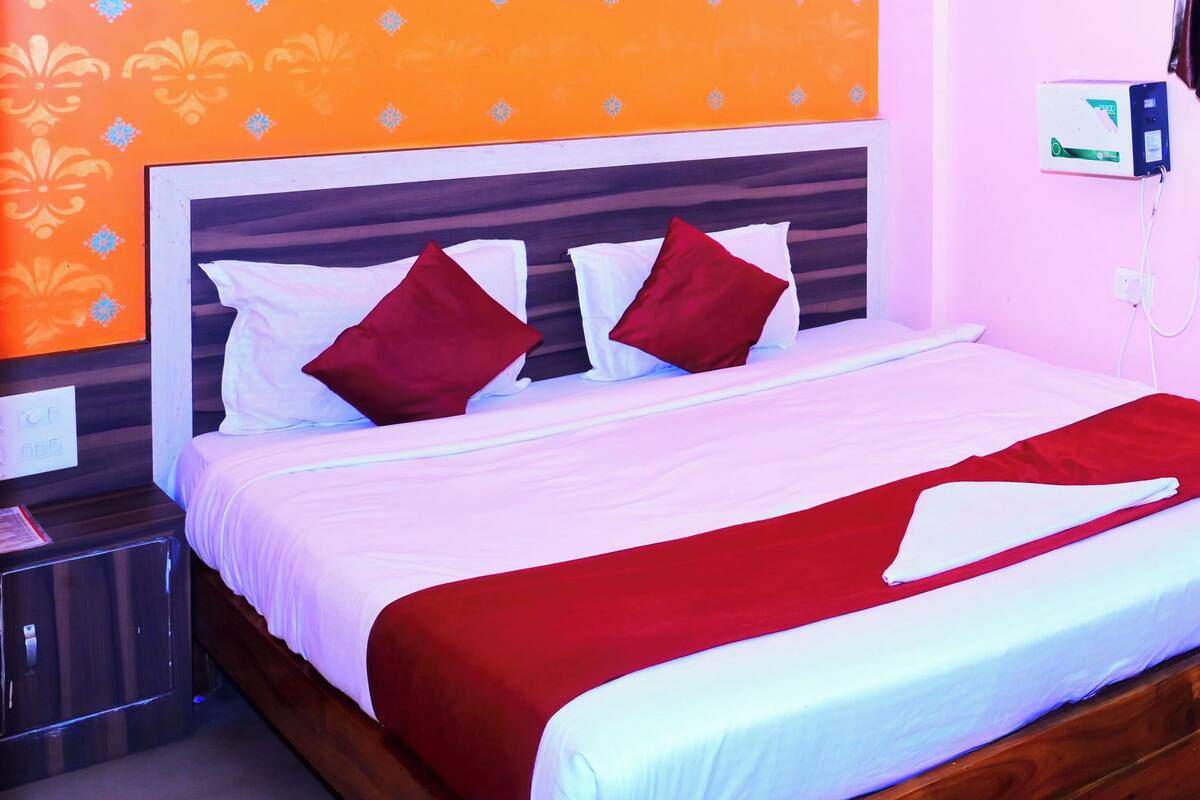 Habitación de hotel con pared de diseño anaranjado, cama con ropa de cama blanca y roja, ventilador de techo y mesita de noche con toma de corriente.