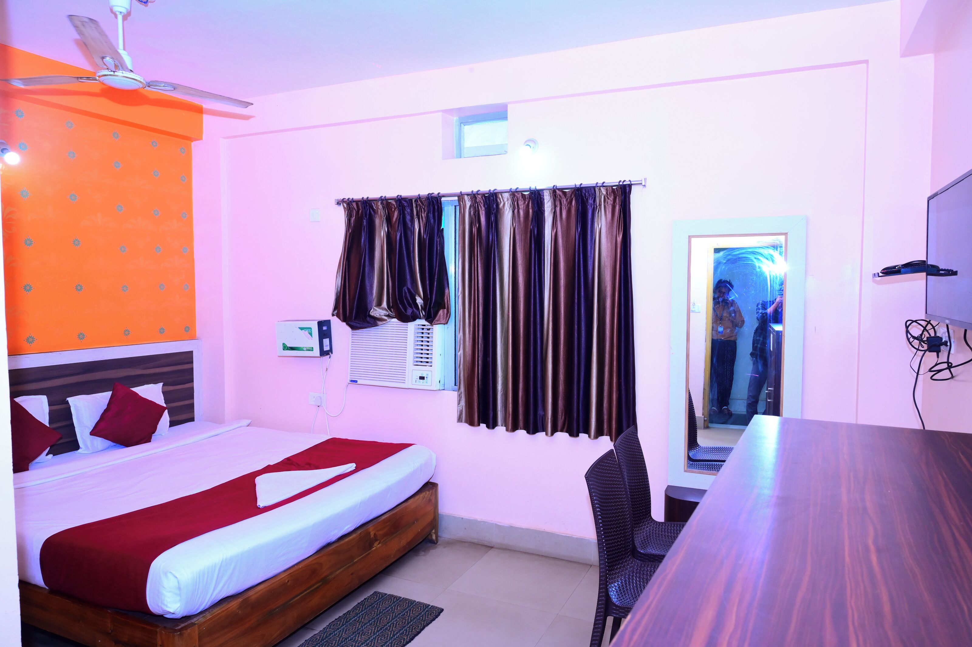 A hotel room with a bed, a desk, and a chair. There is a window with curtains, an air conditioning unit, and a ceiling fan.