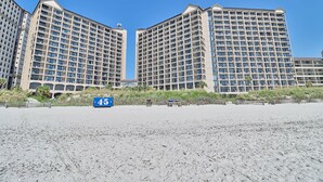 Un hôtel en bord de mer composé de plusieurs immeubles de grande hauteur, avec une plage de sable en premier plan et un ciel bleu dégagé.