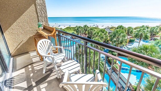 Un balcon avec des chaises blanches, une piscine et une vue sur la plage et l’océan.