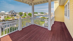 A covered porch with a wooden ceiling and white railings, overlooking a marina with boats and houses.