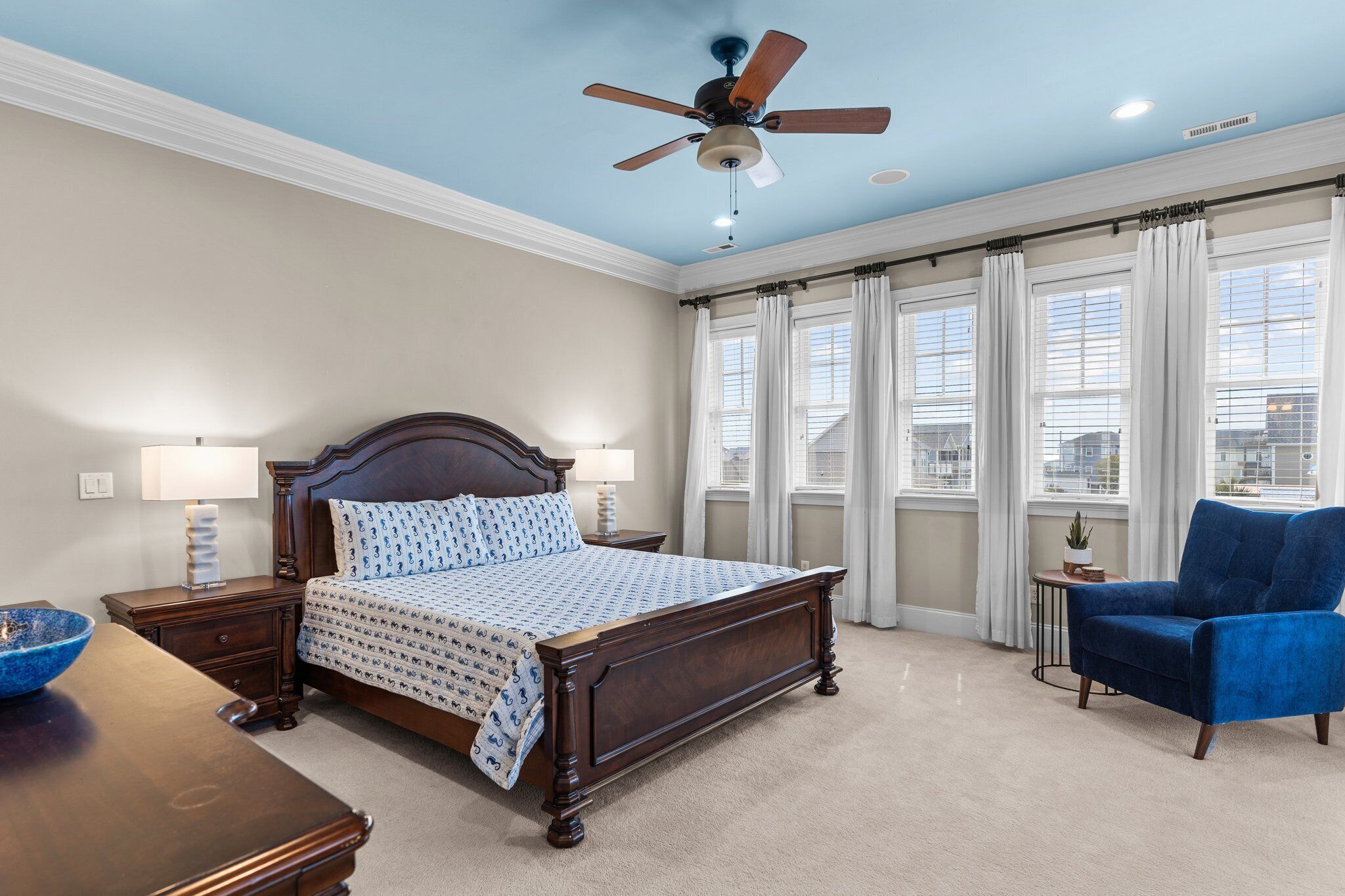 A bedroom with a bed, a ceiling fan, a nightstand, a lamp, a chair, and a window with curtains.