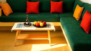 Un sofá verde con cojines rojos y amarillos, una pequeña mesa de centro de madera con una botella y manzanas, y tres cuadros en la pared.