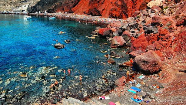 Una zona costera con agua azul cristalina, terreno rocoso y una playa con gente.