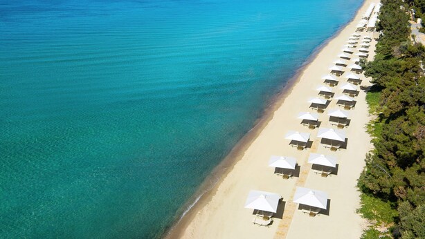 An aerial view of a beach with rows of sunbeds and umbrellas along the shoreline, a clear blue sea, and a distant mountain range. - Chalkidiki Sea Aura - Eva s Cozy Getaway (Nea Propontida)