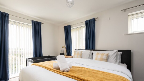 A hotel room with a large bed, a bedside table, a lamp, and a view of the outdoors through the windows. - Spacious 4bd 3.5bt - Etihad COOP -Cinema PoolTable (Greater Manchester)