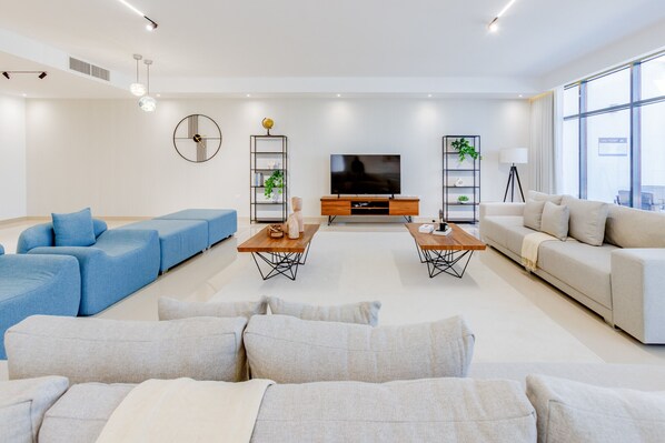 A modern living room with a blue sectional sofa, a grey sofa, and a beige sofa. There are two wooden coffee tables with decorative items. A TV is mounted on the wall, and there are two floor lamps. - Pool, Mountain & Sea View 6BR Villa w/ Asst & Driver Room,Heated Pool,BBQ Area (Fujairah)