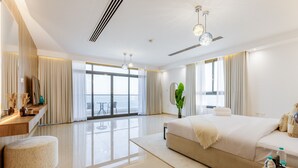 A modern hotel room with a large bed, a sitting area, a vanity with a mirror, and a view of the sea through large windows. - Pool, Mountain & Sea View 6BR Villa w/ Asst & Driver Room,Heated Pool,BBQ Area (Fujairah)