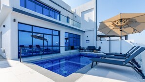 A modern outdoor pool area with a glass balustrade, lounge chairs, and a large umbrella. - Pool, Mountain & Sea View 6BR Villa w/ Asst & Driver Room,Heated Pool,BBQ Area (Fujairah)
