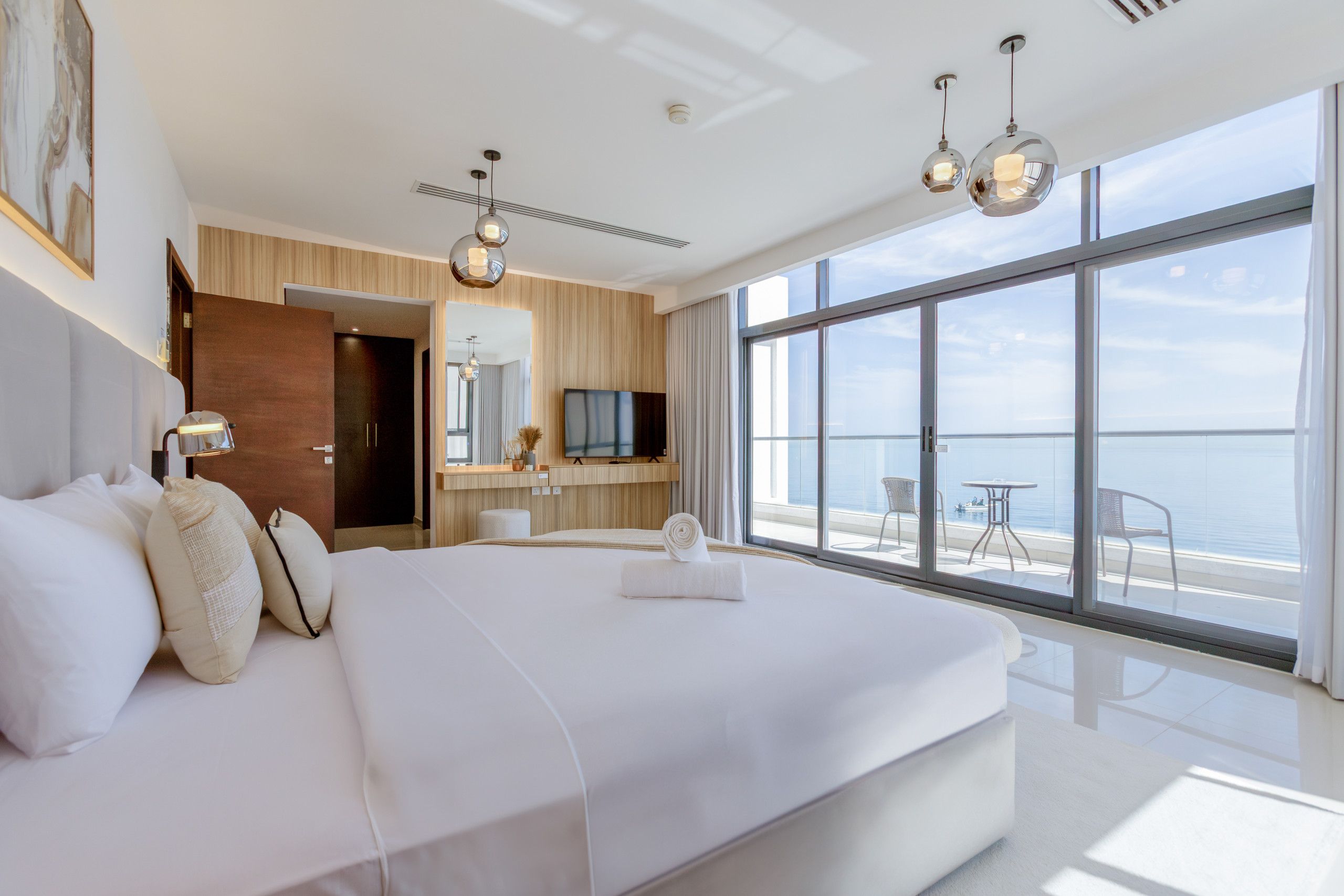 A modern hotel room with a large bed, a desk, and a TV. There is a balcony with a view of the sea.