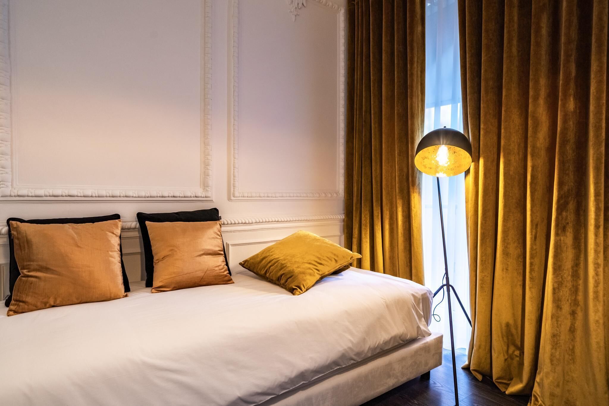 Cama bem arrumada com lençóis brancos e três almofadas douradas, uma luminária de chão e uma janela grande com cortinas.