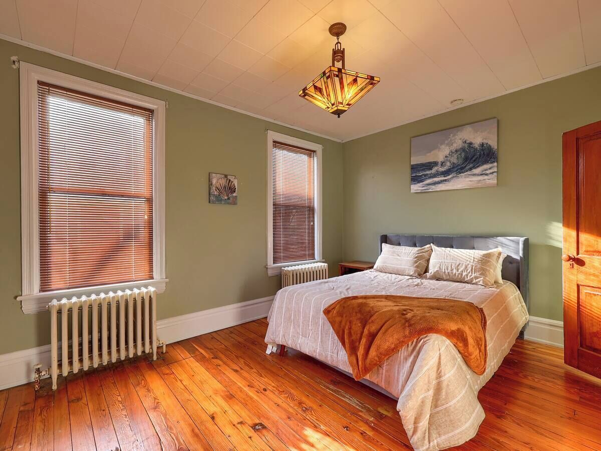 A bedroom with a bed, two windows with blinds, a radiator, a wooden floor, and a painting on the wall.