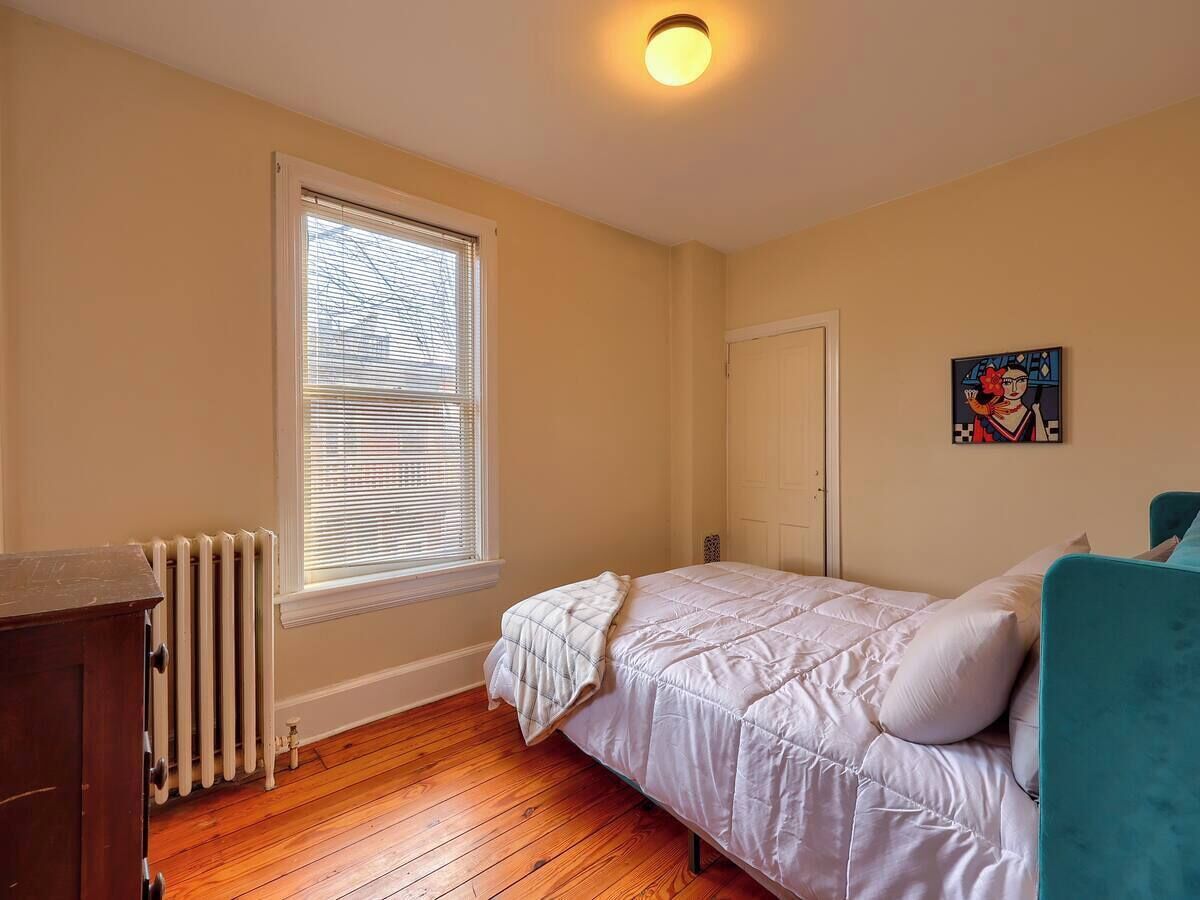 A bedroom with a bed, a radiator, a window with blinds, a door, and a wall-mounted artwork.