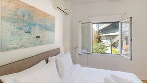 A hotel room with a bed, white linens, a painting on the wall, and a window with a view of buildings and greenery. - Acropolis Museum Retreat by GHH (Athina)
