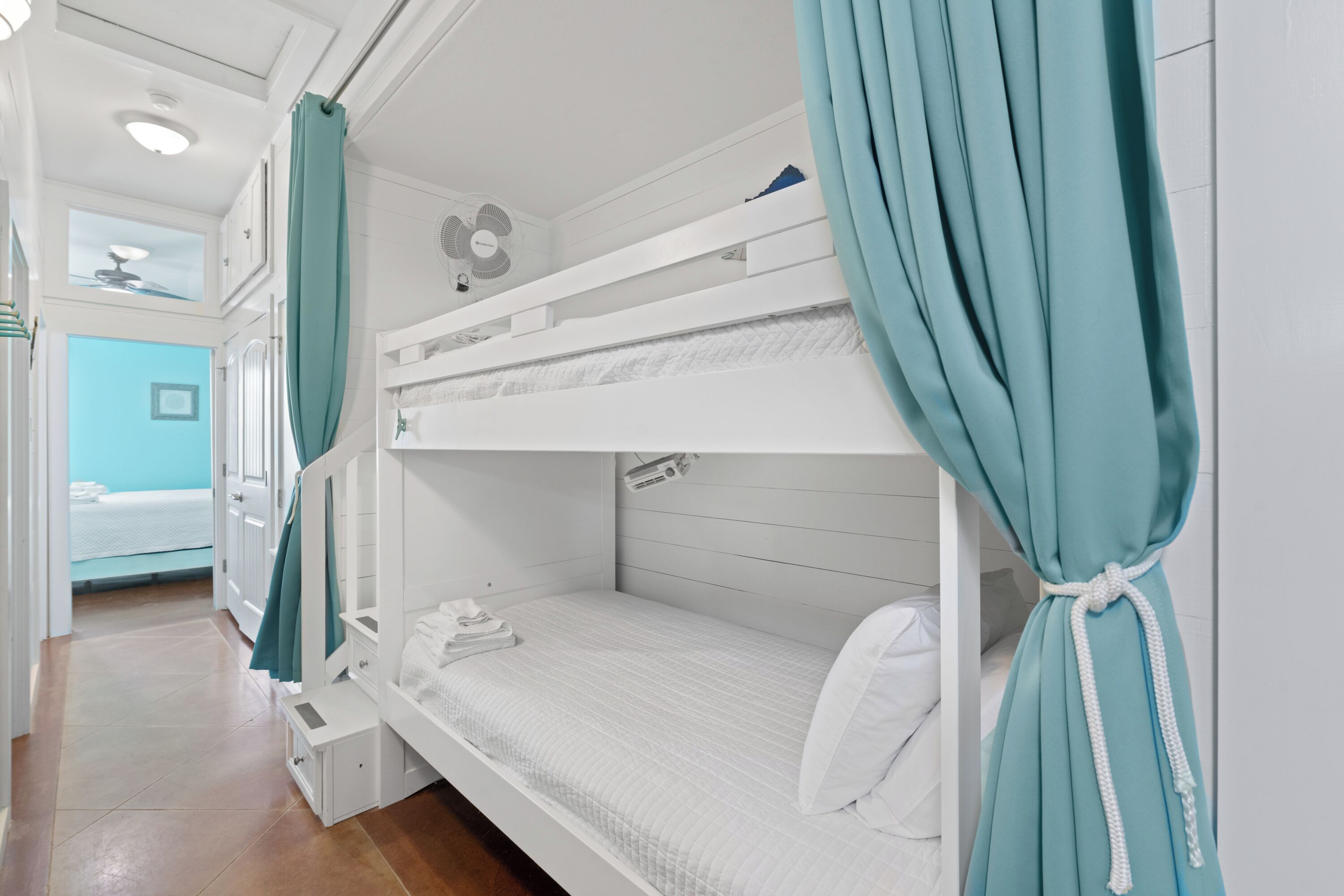 Ein Schlafraum mit Etagenbetten, türkisblauen Vorhängen, einem Deckenventilator und einem kleinen Fenster.