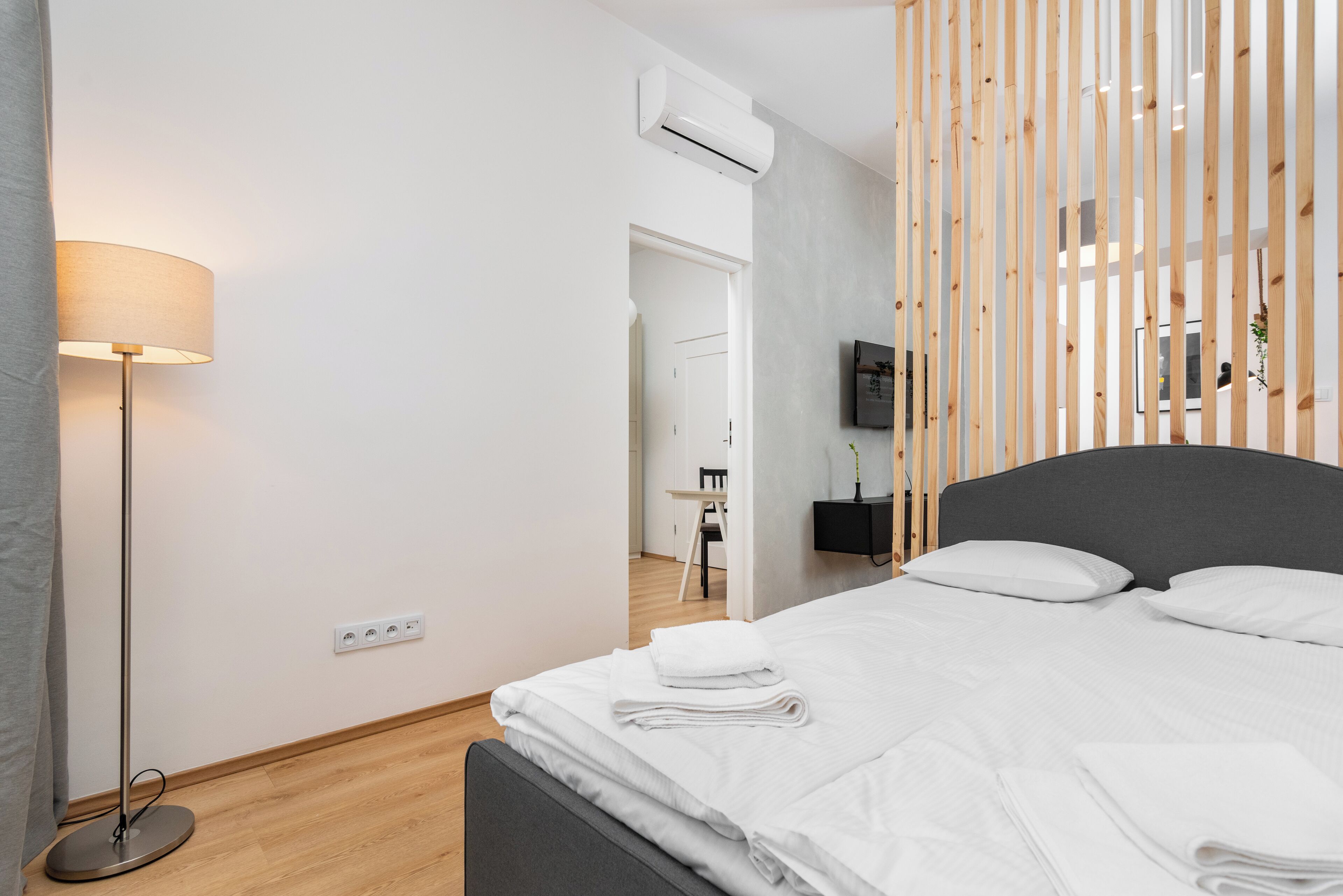A modern hotel room with a neatly made bed, a standing lamp, a flat-screen TV mounted on the wall, and a wooden partition.