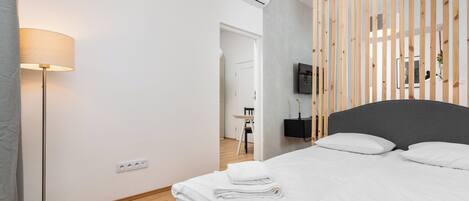 A modern hotel room with a neatly made bed, a standing lamp, a flat-screen TV mounted on the wall, and a wooden partition.