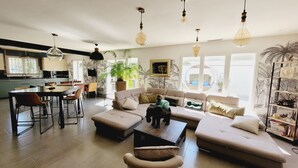 A spacious living area with a sofa, coffee table, and dining table. There is a kitchen in the background with a TV and a plant.