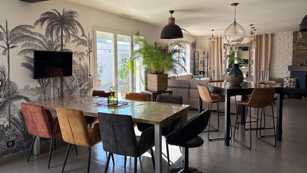 A modern kitchen with a dining area, featuring a large island, chairs, and a TV mounted on the wall.