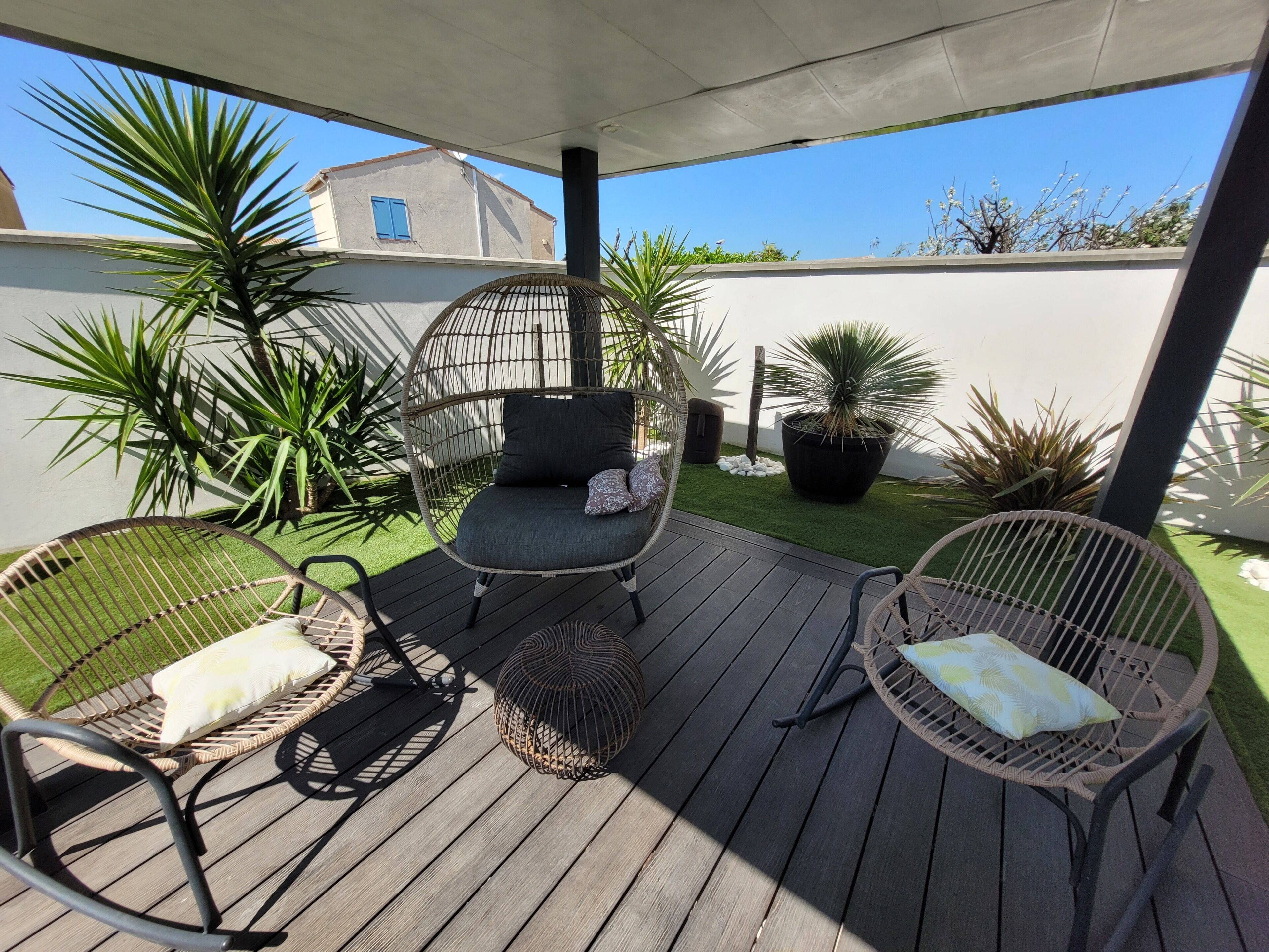 A covered outdoor seating area with wicker chairs and cushions, surrounded by potted plants and a small patch of artificial grass.