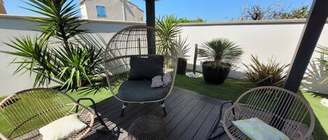 A covered outdoor seating area with wicker chairs and cushions, surrounded by potted plants and a small patch of artificial grass.