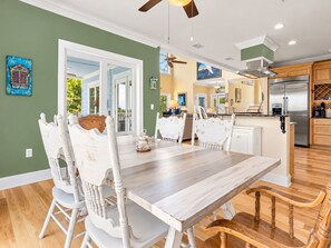 A dining area with a wooden table and white chairs, a kitchen with a stainless steel refrigerator, and a ceiling fan. - Elegant Escape: Pool-Gourmet-Kitchen-Elevator (Long Beach)