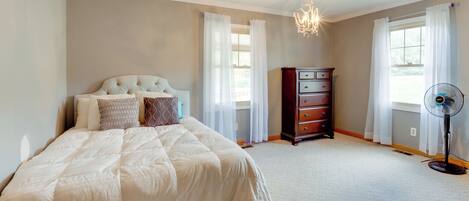 A bedroom with a large bed, a dresser, a fan, and two windows with curtains.
