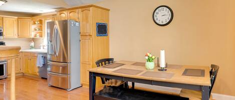 A kitchen with wooden cabinets, a stainless steel refrigerator, a dining table with chairs, and a wall clock.