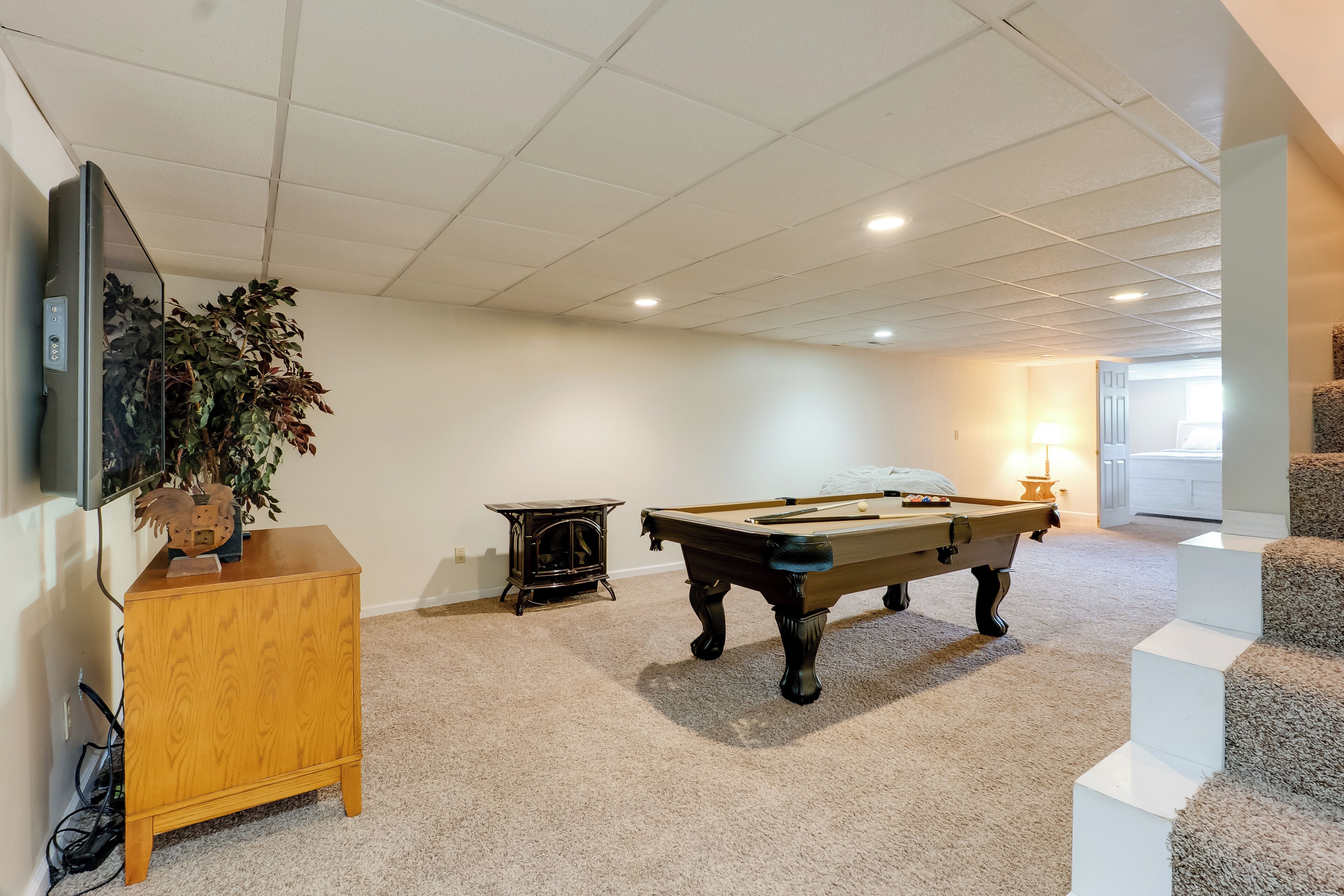 A pool table in a room with carpeted flooring, a wooden cabinet, and a television mounted on the wall.