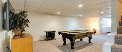 A pool table in a room with carpeted flooring, a wooden cabinet, and a television mounted on the wall.