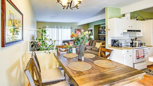 Un coin repas avec une table en bois, des chaises et un lustre. On aperçoit, au fond, une cuisine avec des meubles blancs et une cuisinière.