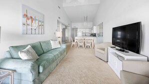 A modern living room with a teal sofa, a flat-screen TV, and a dining area with white chairs.