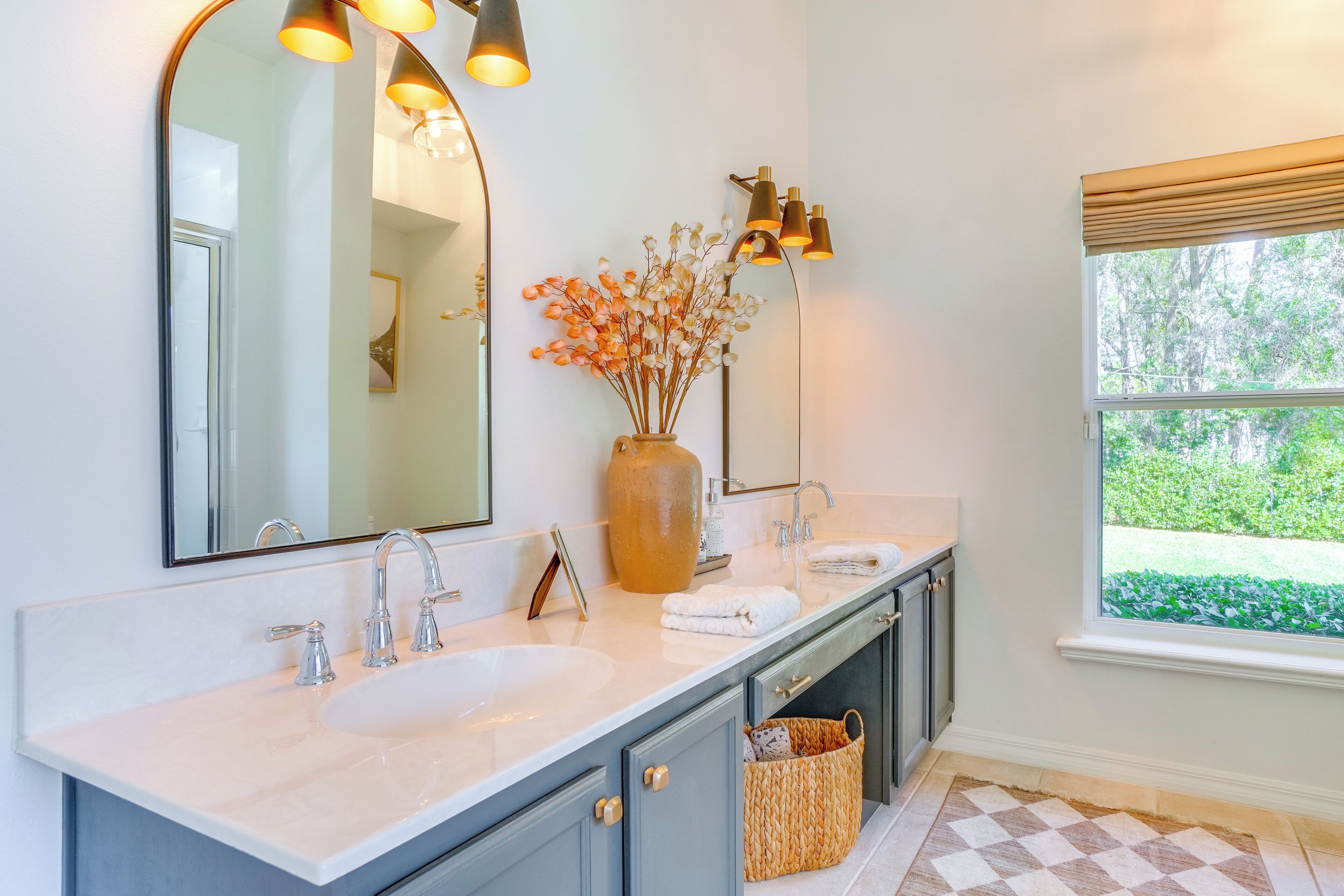 A bathroom with a large mirror, two sinks, a vase with dried flowers, and a window with a view of greenery.